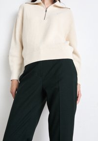 Kremowy sweter z ribbowanym splotem i kołnierzykiem na ćwierć zamka, połączony z ciemnozielonymi spodniami o wysokim stanie, które charakteryzują się prostymi liniami i brakiem widocznych wzorów.