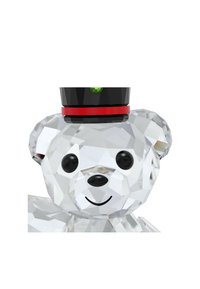 Figúrka medveďa vyrobená z kryštálu s fazetovaným dizajnom, čiernym klobúkom, červeným pásom a akcentom v podobe zelenej drahokamu. Má čierne oči a usmievavý výraz.