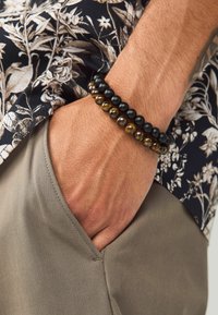 Zwei bezaubernde Armbänder: eines mit glatten schwarzen Perlen und ein anderes mit gemusterten goldenen und braunen Perlen. Getragen an einer Hand mit einem blumigen Hemd.