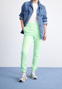 Person som bär ljusgröna joggare, vita sneakers, vit skjorta och blå denimjacka med händerna i fickorna, stående på blått golv.