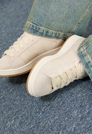 Osoba nosząca beżowe zamszowe sneakersy z kremowymi sznurówkami i jasnoniebieskie dżinsy, stojąca na niebieskim dywanowym podłożu.
