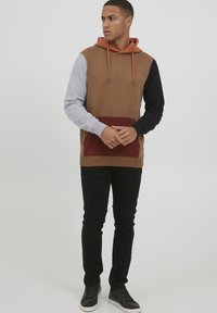 Hooded sweatshirt with brown, gray, black, and burgundy panels. It features a front pocket and drawstrings. Paired with black pants and sneakers.