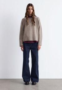 Helles beigefarbenes Wollpullover mit geripptem Saum, über einem dunkelburgunderfarbenen Shirt getragen, kombiniert mit weiten, dunkelblauen Jeans und dunklen Schuhen.