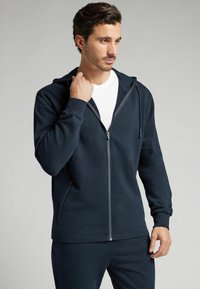 Sudadera con cremallera en color marino, confeccionada en una tela suave, que presenta una cremallera frontal, capucha ajustable con cordón y bolsillos laterales. Diseño liso y casual.