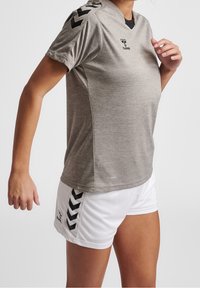 Persona atletica che indossa una maglietta sportiva grigia a maniche corte e pantaloncini bianchi con strisce di logo nere, posando contro uno sfondo semplice.