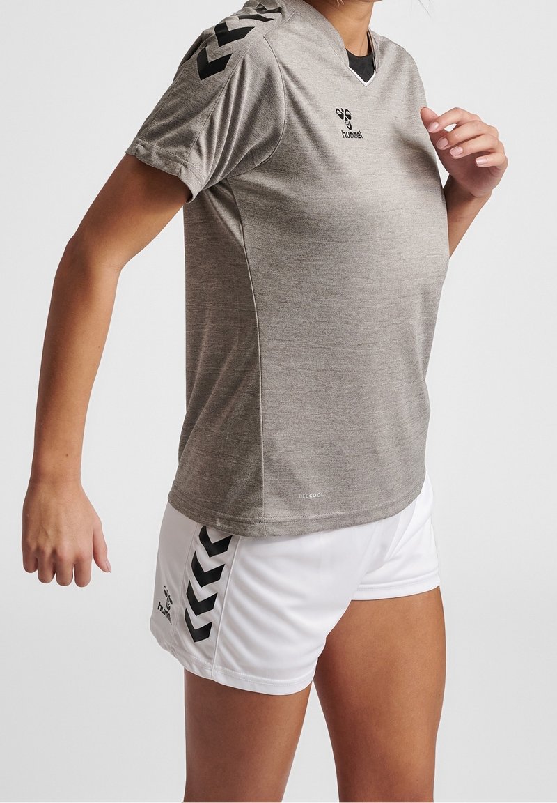 Persona atletica che indossa una maglietta sportiva grigia a maniche corte e pantaloncini bianchi con strisce di logo nere, posando contro uno sfondo semplice.