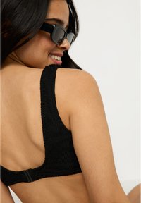 Smaidīga sieviete ar gariem tumšiem matiem, tērpusies melnā teksturētā bikini augšdaļā un melnos saulesbrillēs, skatoties prom pret vienkāršu fona.