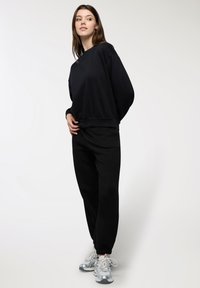 Giovane donna in posa con un grande felpa nera, pantaloni jogger neri e sneakers argentate su uno sfondo chiaro e semplice.