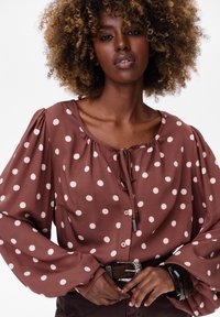 Brązowa bluzka w kropki z okrągłym dekoltem, detalem w postaci wiązania oraz długimi bufkami. Posiada zapięcie na guziki oraz miękką, gładką fakturę materiału.