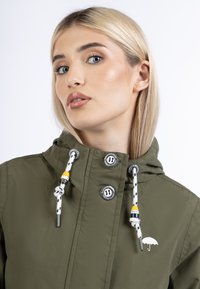 Chaqueta verde oliva con capucha, que presenta botones metálicos, cordones con detalles en amarillo y un logo de paraguas blanco cerca del cuello.