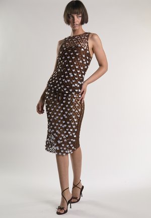 Kobieta o krótkich włosach, ubrana w brązową, bezrękawnikową sukienkę z siateczki ozdobioną dużymi białymi cekinami, pozująca w czarnych sandałkach na wysokim obcasie.