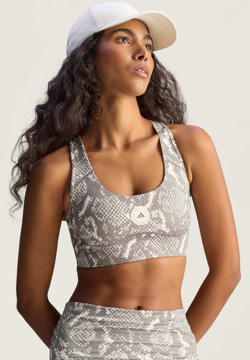 Vrouw met lang krullend haar, gekleed in een lichte sportbeha met slangenprint, bijpassende leggings en een witte baseballpet, die wegkijkt.