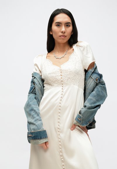 Šviesaus dramblio kaulo spalvos satino suknelė su nėrinių liemenėle, puošta sagų detalėmis priekyje; derinama su nusidėvėjusia džinsine striuke kontrastui.