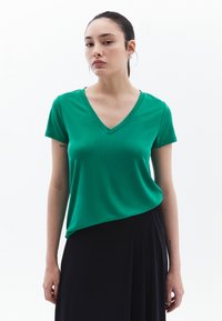 Camiseta verde de manga corta con cuello en V en tejido suave, con un diseño ajustado y un dobladillo asimétrico, que se combina con una falda negra.