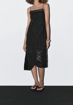 Vrouw draagt een mouwloze, zwarte kanten midi-jurk met dunne bandjes en zwarte sandalen, staand tegen een effen witte achtergrond.