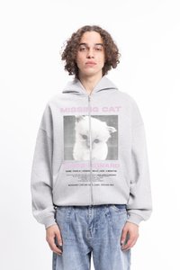 Grå hettegenser med glidelås som har et grafisk bilde av en savnet katt-plakat, med svart og rosa tekst. Kombinert med løse, lyseblå denimbukser.