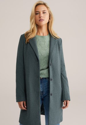 Groene wollen blend jas met een geknipt revers, twee knopen en lange mouwen, gelaagd over een gestructureerd lichtgroen sweater en een blauwe spijkerbroek.