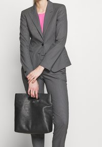 Costume gris sur mesure avec un haut rose, associé à un sac cabas en cuir noir avec une texture lisse et des poignées découpées.