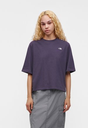 Jonge vrouw met lang blond krullend haar, draagt een los donkerpaars T-shirt en een grijze cargorok, staand tegen een effen achtergrond.