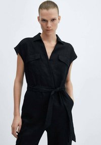 Vrouw in een zwart, mouwloos jumpsuit met ceintuur en borstzakken, staand tegen een effen lichte achtergrond.