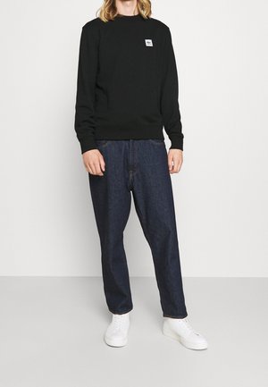 Felpa nera in cotone con polsini e orlo a coste, caratterizzata da una piccola etichetta con logo bianco. Abbinata a jeans in denim scuri larghi e sneakers bianche.