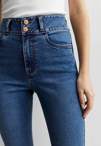 Jeans de mezclilla azul de cintura alta con dos botones, detalles de costura sutil y bolsillos estándar a ambos lados. Textura suave.