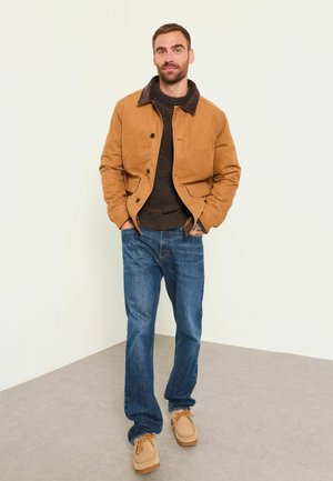 Mann steht mit Händen in den Taschen, trägt eine beige Jacke, einen dunklen Pullover, blaue Jeans und beige Schuhe vor einer schlichten weißen Wand.