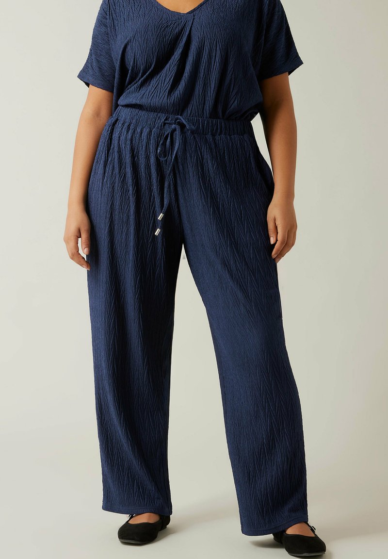 Femme portant un haut texturé bleu marine à manches courtes et un pantalon large assorti avec un cordon de serrage, debout les mains détendues le long du corps.