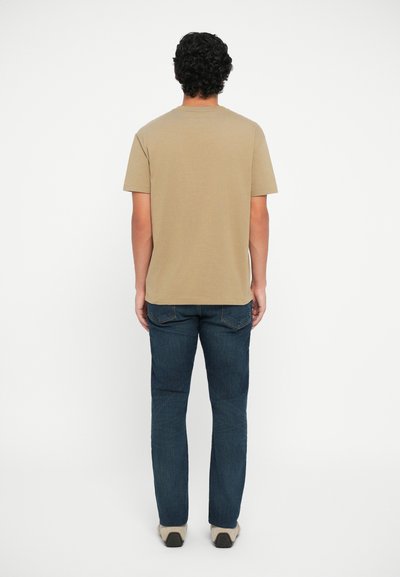 Tricou din bumbac bej, cu mâneci scurte, văzut din spate, asortat cu blugi albaștri și pantofi de culoare deschisă. Textură netedă și croială clasică.