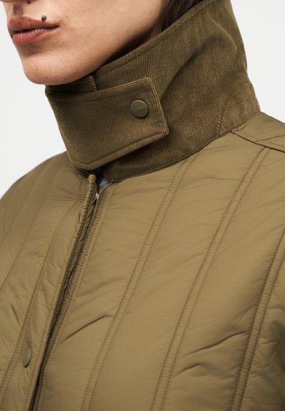 Κοντινή λήψη ατόμου που φοράει μπουφάν ελιάς με καπιτονέ ύφασμα, ψηλό γιακά από κορντούρα και κούμπωμα με κουμπιά σκράτς.