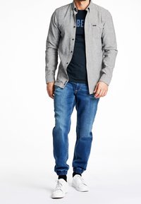 Camicia grigia con bottoni sopra una maglietta blu navy, abbinata a jeans blu e sneakers bianche. La camicia ha una tasca; i jeans sono leggermente affusolati.
