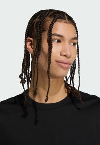 Sort t-shirt på en person med langt, fletet hår, der har små sektioner sikret med sorte elastikker og en glat tekstur. Baggrunden er lys.