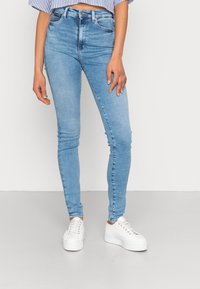 Mujer vestida con jeans ajustados de color azul claro y zapatillas blancas de plataforma, de pie contra un fondo liso.