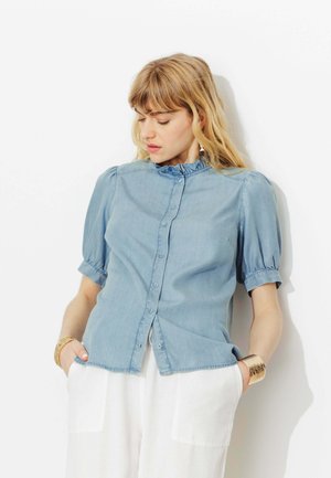 Femme aux cheveux blonds portant un chemisier bleu clair boutonné et un pantalon blanc, debout les mains dans les poches devant un fond blanc.