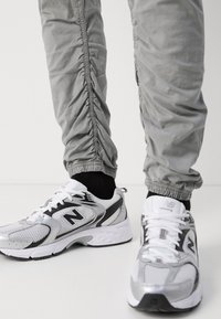 Szare elastyczne spodnie z marszczeniami po bokach zestawione z białymi i srebrnymi butami do biegania New Balance, które mają akcenty z siateczki i gumy.