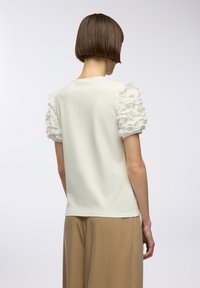 Top bianco con maniche a volant in tessuto trasparente, scollo rotondo semplice e texture liscia. Abbinato a pantaloni beige a gamba larga.