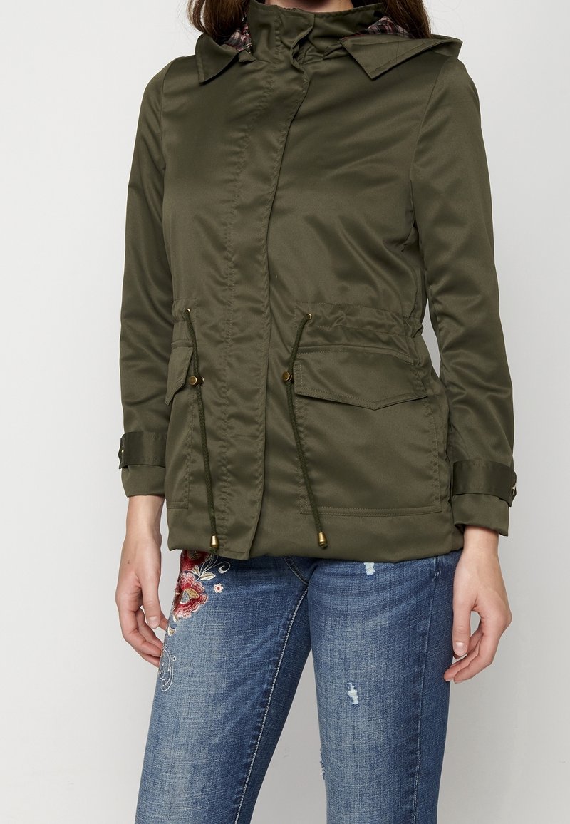 Kobieta w oliwkowej, długiej kurtce z kapturem, z przednimi kieszeniami i sznurkiem, w połączeniu z niebieskimi, wyszywanymi dżinsami zniszczonymi.