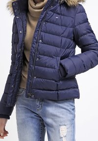 Osoba v námořnickém modrém péřovém kabátu s kapucí lemovanou kožešinou, béžovém roláku a světle modrých džínách s dírami, s jednou rukou v kapse.
