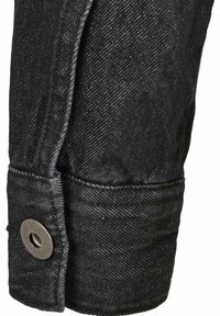 Svart denimjacka med detaljer som en uppvikt ärm, metalltryckknapp och en texturerad yta med ett subtilt diagonalt vävmönster.