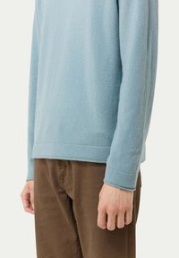 Hellblauer Pullover mit langen Ärmeln aus weichem Material, ausgestattet mit gerippten Bündchen und Saum. Kombiniert mit braunen Hosen, die die lockere Passform betonen.