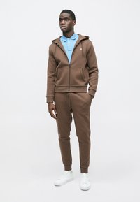 Brown zip-up hoodie and matching joggers made of soft fabric, worn over a light blue polo. Accented with a small logo on the chest. White sneakers.