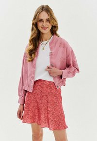 Růžová zkrácená bunda s květinovou výšivkou, spárovaná s červenou sukní s květinovým potiskem. Model má na sobě bílé tričko a vrstvené náhrdelníky.