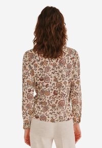 Langærmet bluse i lys beige med et blomstermønster i rød, orange og grøn. Har en tætsiddende design og knapdetaljer ved manchetterne.