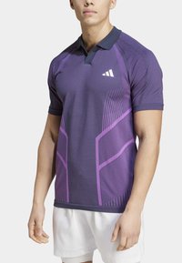 Camiseta polo morada con un cuello oscuro en contraste, que presenta líneas diagonales audaces y un logo blanco en el pecho. Manga corta, ajuste atlético.