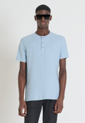Camisa henley de manga corta en azul claro con un acabado texturizado, con una tapeta de tres botones y un pequeño logo bordado. Se lleva con gafas de sol negras.