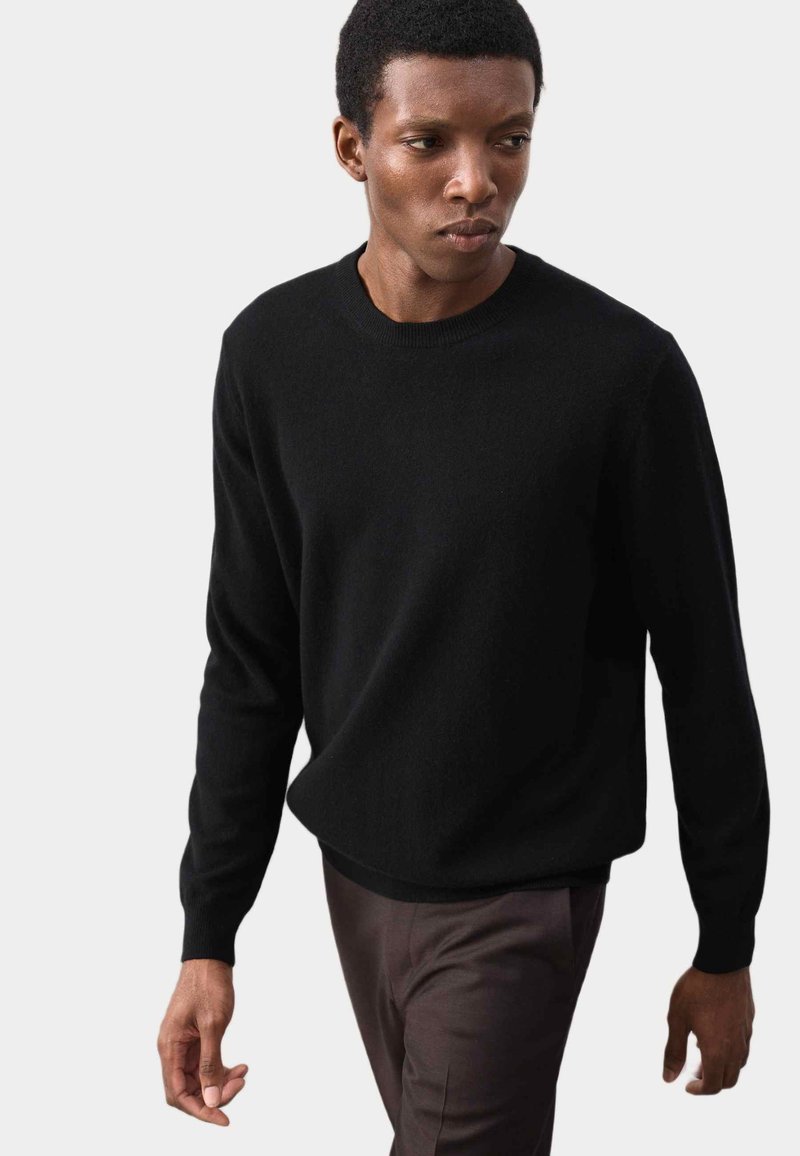 Pull en maille noire avec un col rond et des manches longues, associé à un pantalon marron foncé ; texture lisse et design minimaliste.