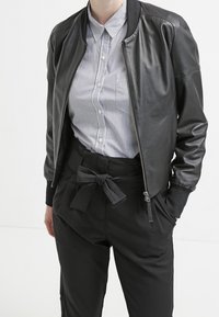 Veste aviateur en cuir noir avec poignets et col côtelés, fermeture éclair à l'avant, associée à une chemise rayée à boutons et un pantalon noir taille haute.