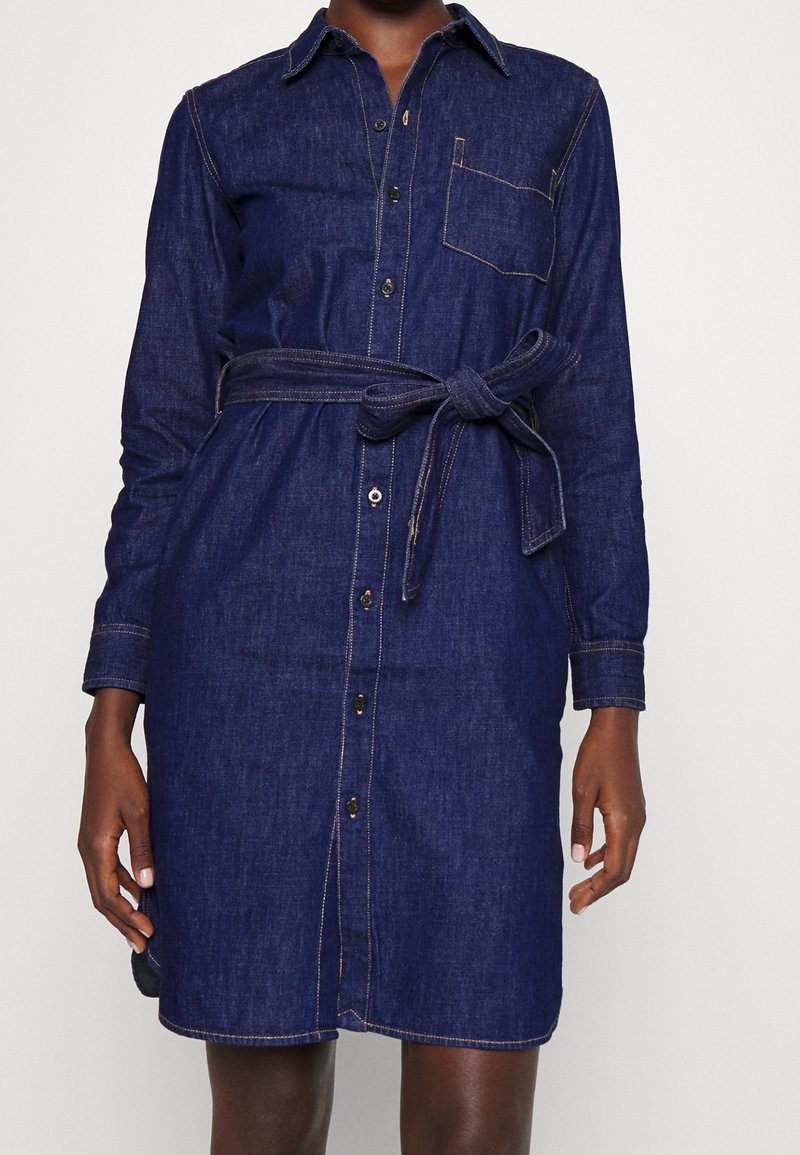 Kvinna som bär en långärmad, mörkblå denimklänning med knäppning framtill, bröstficka och knytbart midjebälte.