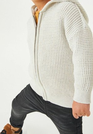 Kind trägt eine cremefarbene Strickjacke mit Reißverschluss, schwarze Hose und braune Schuhe und macht einen Schritt nach vorne vor weißem Hintergrund.