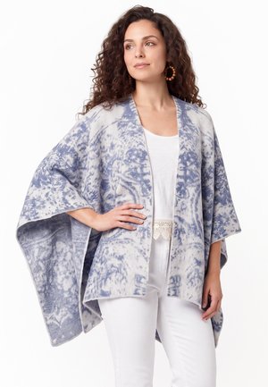 Frau mit lockigem Haar trägt einen blau-weiß gemusterten Poncho über einem weißen Top und einer weißen Hose, steht mit einer Hand in der Hüfte.